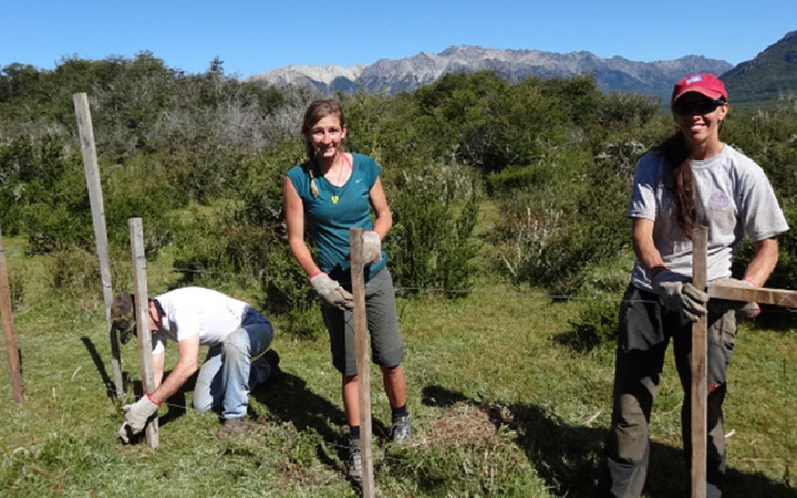 Three people work on fence posts during a service project with outward bound. There are mountains in the background. 